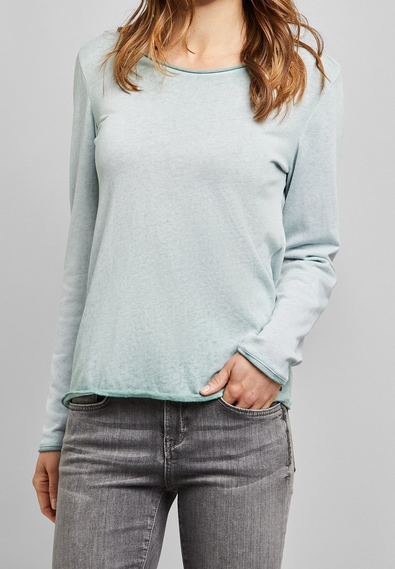 Femme portant une chemise à manches longues bleu clair et un jean gris, une main dans la poche, sur un fond neutre.
