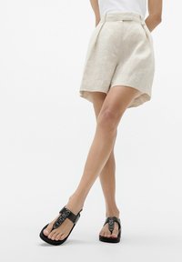 Pantaloni corti in lino beige chiaro con vestibilità ampia e pieghe. Sandali neri con una fascia testurizzata e suola piatta, dal design casual.