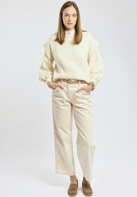 Femme portant un pull sans manches en maille torsadée crème sur une blouse à manches ballon, un pantalon large crème, des mocassins à imprimé léopard et un bracelet doré.