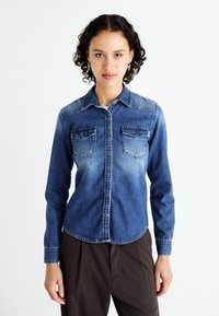 Camicia di jeans blu scuro con due tasche sul petto, chiusura con bottoni, colletto a punta e orlo curvo. Presenta zone sbiadite e decorazioni cucite.