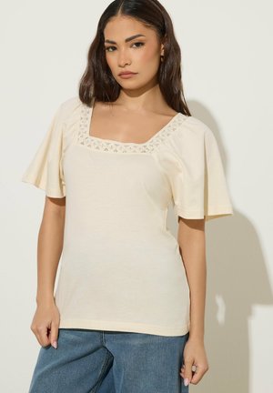 Jonge vrouw met lang donker haar, gekleed in een crèmekleurige korte mouw top met kanten vierkante halslijn en blauwe spijkerbroek, staand tegen een beige achtergrond.