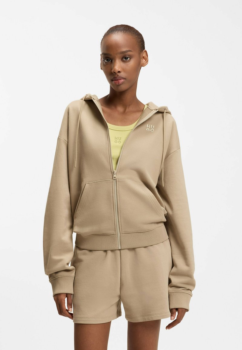 Frau trägt eine beige Zip-Hoodie und passende Shorts über einem hellgrünen Top mit "HUGO"-Logo auf beiden Kleidungsstücken, steht vor weißem Hintergrund.