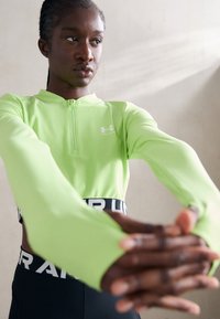 Hosszú ujjú, félcipzáros sportfelső élénk lime zöld színben, magas gallérral, testhezálló fazonnal és fekete derékpánttal, amelyen merész fehér betűk találhatók.