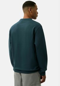 Sudadera de un tono verde azulado oscuro con un cuello redondo y mangas largas, que presenta un corte holgado y puños de canalé. Vista desde la parte posterior.