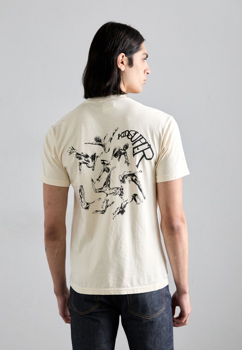 Tricou crem cu mânecă scurtă, având un imprimeu grafic negru pe spate cu figuri dinamice și cuvântul "KID SUPERT" într-un design arcuit.