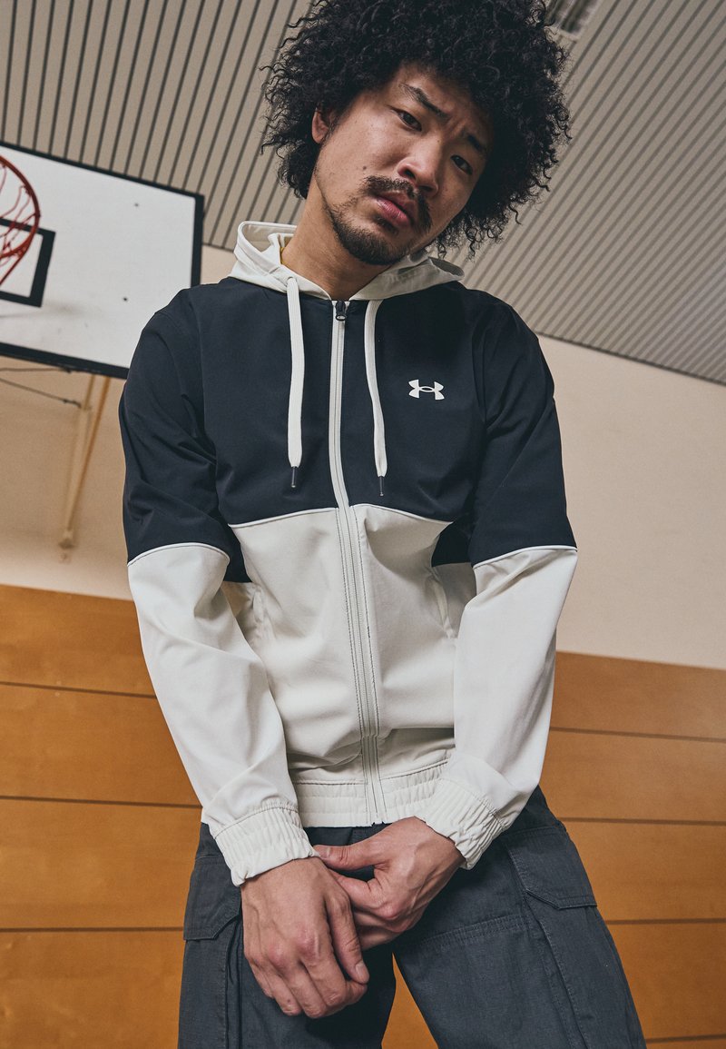 Giacca nera e bianca con zip, polsini elasticizzati e cappuccio. Presenta un logo Under Armour sul petto. Il modello indossa pantaloni cargo grigi.