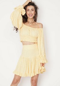 Żółty top w stylu off-shoulder z falbaniastymi rękawami i dopasowaną spódnicą w kształcie dzwonu. Posiada kwiatowe wzory i marszczoną strukturę.