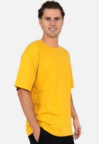 T-shirt en coton jaune avec des manches courtes, un col rond et une coupe décontractée. Porté avec les mains dans les poches et un pantalon noir uni.