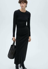 Robe noire à manches longues et ajustée avec un col rond et un drapage sur les côtés. Associée à un sac fourre-tout noir structuré et des bottes à talons épais.