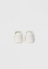 Baskets blanches avec une finition en cuir lisse, un design à bout rond, une semelle plate et un logo au talon. Apparence minimaliste sans motifs.