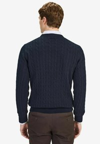 Laivastonsininen kaapelikudottu neulepusero, jossa on ribbireunat hihoissa ja vyötäröllä. Siinä on korkea kaulus ja teksturoitu kuvio. Pusero on puettu kauluspuseron päälle.