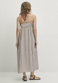 Długa sukienka na ramiączkach, z marszczoną górą i swobodnym dołem w jasnobrązowym, teksturowanym materiale. Całość dopełniają czarne, płaskie sandały.