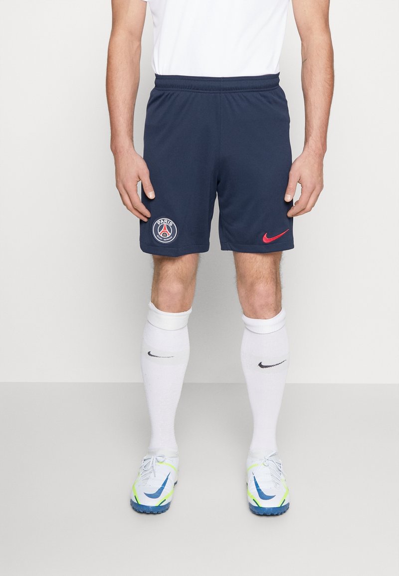 Atleta masculino vistiendo pantalones cortos deportivos azul marino del Paris Saint-Germain, calcetas blancas hasta la rodilla con logotipos negros de Nike y zapatos de fútbol blancos con detalles en azul y neón.
