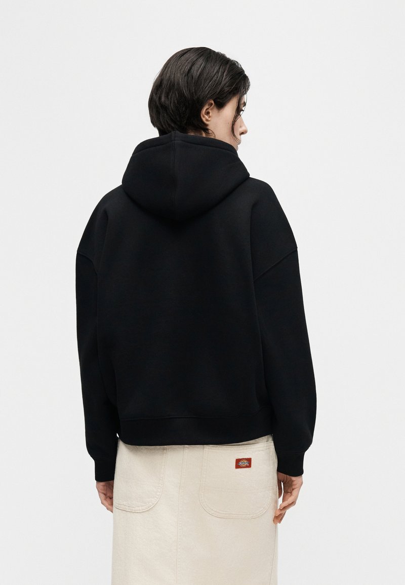 Person with short dark hair, wearing a black hooded sweatshirt and a light-colored skirt with large back pockets, standing against a plain background.