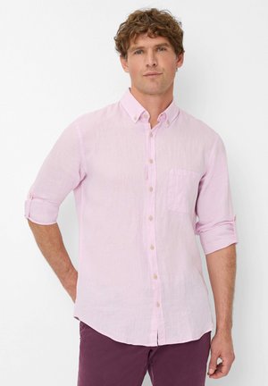 Camicia rosa chiaro con bottone in tessuto leggero; presenta maniche arrotolabili, una singola tasca sul petto e una trama sottile. Abbinate a pantaloni viola scuro.