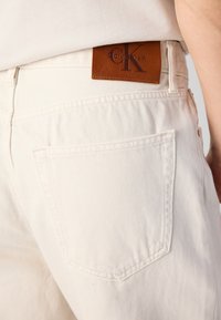 Close-up of light beige jeans back pocket with brown Calvin Klein leather brand patch on waistband.