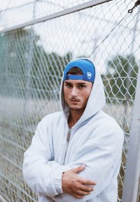 Siva zip-up dukserica s prevelikim rukavima, nošena preko plave bejzbol kape pričvršćene straga, protiv pozadine ograde od lanca.