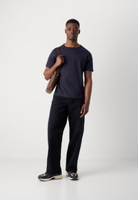 Marineblaues T-Shirt im lässigen Schnitt, dunkle Jeans und mehrfarbige Sneakers. Braune Tasche über einer Schulter. Glattes Material, lässiges Design.