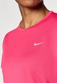 Tricou sportiv roz, fabricat dintr-un material neted. Dispune de un guler rotund și un mic logo Nike argintiu pe față, conceput pentru confort.