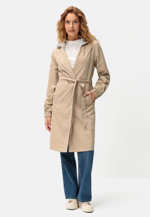 Frau trägt einen beigen, taillierten Trenchcoat über einem weißen Lochmuster-Top, blauen Jeans und beige Sneakern und steht mit einer Hand in der Manteltasche.