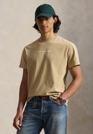 Νέος άντρας που φοράει μπεζ μπλουζάκι Polo Ralph Lauren, πράσινο καπέλο μπέιζμπολ, μπλε τζιν και ρολόι χειρός, με το χέρι στην τσέπη μπροστά σε απλό φόντο.