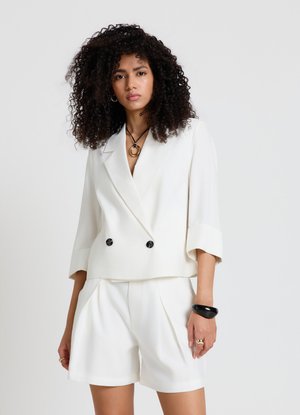 Donna con capelli ricci che indossa un blazer bianco doppiopetto con maniche arrotolate e pantaloncini bianchi coordinati, accessoriata con gioielli neri e dorati.