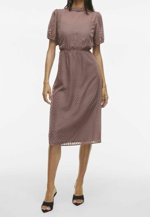 Femme portant une robe mauve longueur genou avec des manches bouffantes et un motif chevron texturé, associée à des talons noirs ouverts à l’avant.