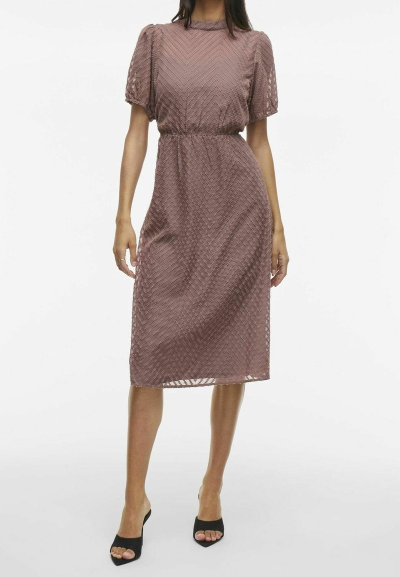 Femme portant une robe mauve longueur genou avec des manches bouffantes et un motif chevron texturé, associée à des talons noirs ouverts à l’avant.