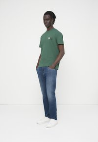 Νεαρός άνδρας με σκούρο δέρμα και κοτσίδες στέκεται με τα χέρια στις τσέπες, φορώντας πράσινο T-shirt, μπλε τζιν και λευκά αθλητικά παπούτσια, σε λευκό φόντο.