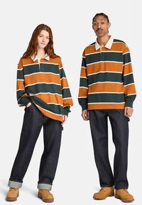 Twee personen dragen gestreepte poloshirts met lange mouwen in oranje en groen, gecombineerd met donkere spijkerbroeken en beige werkschoenen.