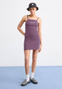 Vestido de alças riscado com faixas finas, predominantemente em roxo e vermelho. Combinado com um chapéu estilo balde preto, meias tipo crew e sapatos slip-on.