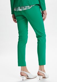 Zielone dopasowane spodnie o wąskim kroju, wykonane z gładkiego materiału, w połączeniu z białymi szpilkami na odkrytej pięcie. Wzorzysta bluzka wystaje spod żakietu.