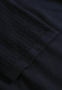 Sötét tengerészkék kötött anyag bordázott és texturált mintákkal, amelyek téglalapalakú részt alkotnak sima kötött háttéren.