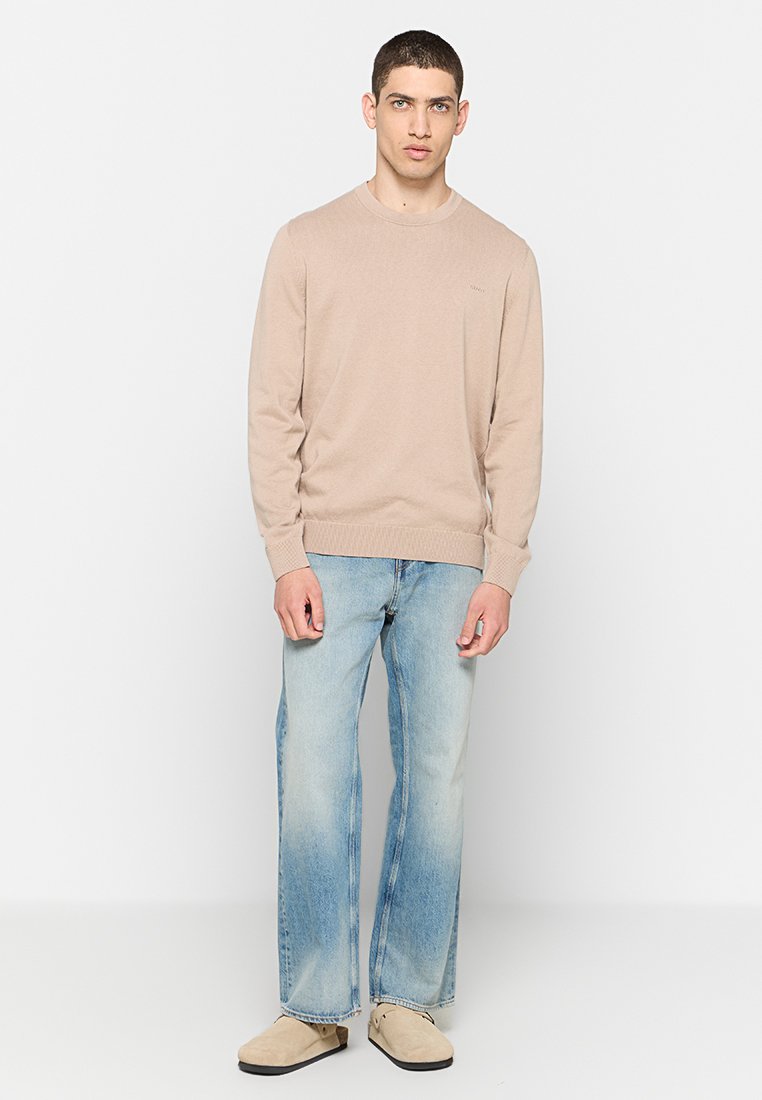 Man staat gekleed in een beige trui, lichtblauwe rechtlopende jeans en beige instappers tegen een witte achtergrond.