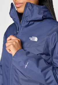 Tummansininen vedenpitävä takki, jossa on korkea kaulus, vetoketjusuljenta ja "The North Face" -logo. Huomattava "DryVent" -merkki hihassa.