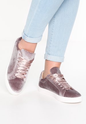 Personne portant un jean bleu clair retroussé et des baskets en velours mauve avec des lacets en satin brillant sur fond blanc.