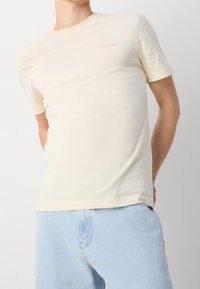 Camiseta de algodón color crema con cuello redondo y mangas cortas. Presenta un discreto logo en marrón claro "Calvin Klein" en el pecho.
