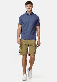 Lachende man die loopt, gekleed in een blauw poloshirt, khaki cargoshorts met zijzakken en witte sneakers, tegen een effen witte achtergrond.