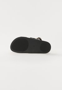 Sandalia negra con suela de goma texturizada, correa ajustable y acento de hebilla metálica. Material superior suave. Forma diseñada para la comodidad.