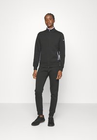 Sudadera negra con cremallera y cuello alto, con rayas laterales blancas en contraste. Combinada con pantalones de chándal negros estrechos y zapatillas deportivas negras.