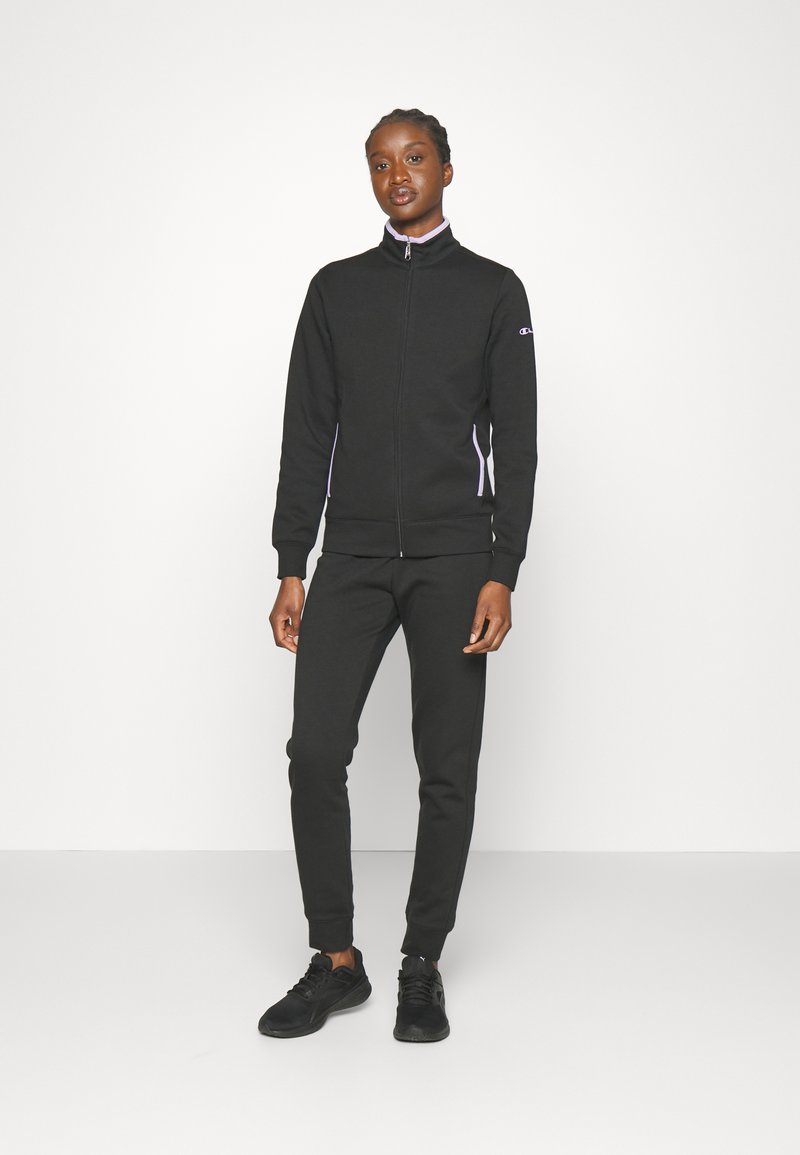 Sudadera negra con cremallera y cuello alto, con rayas laterales blancas en contraste. Combinada con pantalones de chándal negros estrechos y zapatillas deportivas negras.
