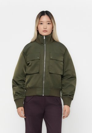 Giovane che indossa una giacca bomber verde oliva con tasche anteriori e pantaloni viola scuro, in piedi davanti a uno sfondo bianco.