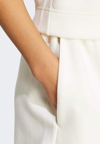 Krémová textília odevu s hladkou štruktúrou, s bočnou vreckom, kde je umiestnená ruka. Šitie je viditeľné blízko pásu.