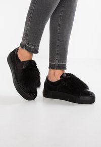 Schwarze Sneakers mit strukturierter Oberfläche, ausgestattet mit einem Überzug aus Kunstfell und einer Schleifenakzent, kombiniert mit grauen cropped Jeans.
