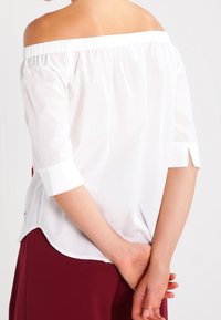 Femme les bras croisés derrière le dos, portant un chemisier blanc à épaules dénudées avec des manches trois-quarts et une jupe bordeaux.