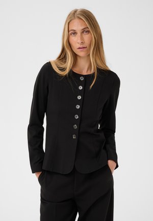 Femme blonde portant une veste noire cintrée boutonnée et un pantalon noir, les mains dans les poches, sur un fond clair uni.