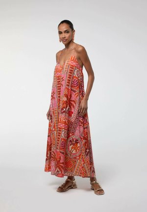 Mujer con el pelo recogido, vestida con un vestido maxi sin mangas, colorido y estampado, y sandalias doradas, de pie frente a un fondo claro y liso.