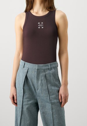 Jeune femme portant un haut sans manches marron foncé avec le logo "JOOP!", associé à un pantalon plissé gris taille haute sur un fond uni.