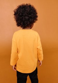 Gul langærmet skjorte med afslappet pasform, båret af et barn. Baggrunden har en matchende orange nuance. Sorte bukser er synlige. Krøllet hår ses bagfra.