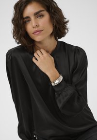 Bluză din satin negru, cu gât înalt și mâneci bufante. Modelul poartă o brățară argintie pe încheietură, evidențiind texturi netede și un design simplu.